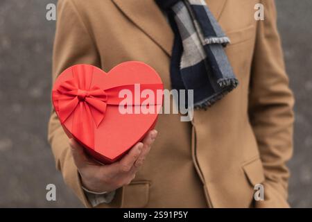 L'uomo felice regala un regalo rosso a forma di cuore con un cappotto beige a San Valentino. Primo piano. Concetto d'amore per il compleanno o l'anniversario. Biglietto di San Valentino. Foto Stock