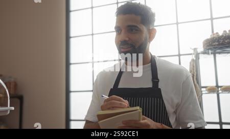 Bel giovane in panetteria che prende appunti con una penna che indossa un grembiule mentre sorride in un ambiente interno Foto Stock
