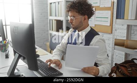 Giovane ispanico che lavora a una scrivania in un ufficio, con occhiali e cravatta, si concentra sullo schermo di un computer mentre tiene in mano documenti, circondato da un'area separata Foto Stock