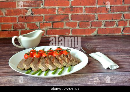 Manzo cotto a pezzi con pomodori ciliegini e asparagi bolliti su un tavolo di legno, accanto a una barca con sugo di carne e posate in un tovagliolo. Primo piano. Foto Stock