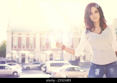 Bella giovane donna alla moda in piedi di fronte a uno sfondo urbano con una tazza usa e getta di caffè caldo Foto Stock
