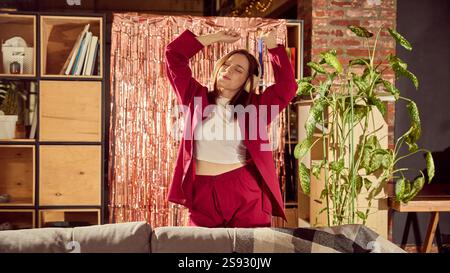 Una giovane donna allegra e rilassata in un ambiente accogliente si indossa ascoltando musica in cuffia, cantando e ballando in un elegante salotto Foto Stock