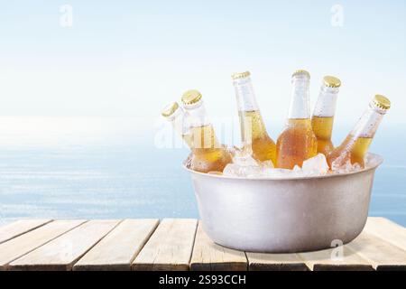 bottiglie fredde di birra in secchio con ghiaccio su tavola di legno sullo sfondo del mare Foto Stock