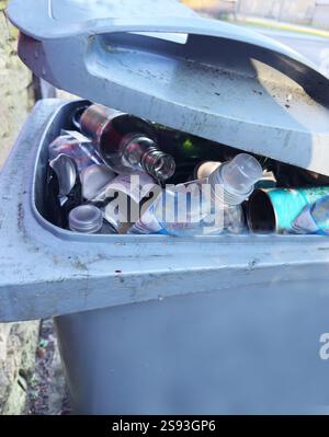 Un contenitore esterno per rifiuti con coperchio che non può chiudersi, poiché il contenitore è pieno di rifiuti riciclabili di bottiglie di plastica e vetro e lattine di latta. Foto Stock