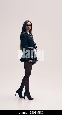 Immagine a figura intera di una bella donna che posa su una giacca corta e una camicia con cravatta e scarpe con tacco sullo sfondo dello studio Foto Stock