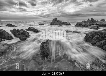 Un primo piano grandangolare di Un'onda che scorre a riva sulle Sea Rocks in Una spettacolare immagine a contrasto elevato in bianco e nero Foto Stock