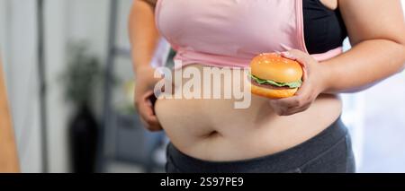 Donna in sovrappeso che tiene in mano un hamburger mentre rivela la sua intersezione, rappresentando sfide di perdita di peso nel viaggio Foto Stock
