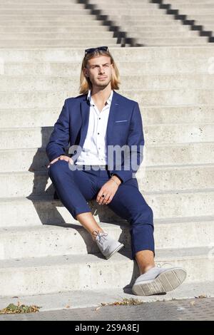 Un giovane con lunghi capelli biondi, con un elegante abito blu e una camicia bianca, posa con sicurezza all'aperto davanti a un moderno schienale architettonico Foto Stock