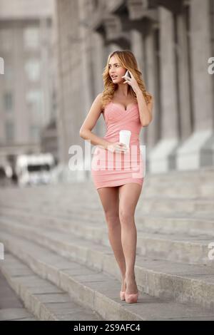 Una bella donna con un vestito rosa che parla al telefono cellulare e tiene una tazza di caffè Foto Stock