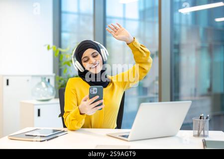 Donna allegra con le cuffie e con il telefono in mano che ascolta musica e guarda video musicali. Donna d'affari in hijab che lavora all'interno dell'ufficio con un computer portatile. Foto Stock