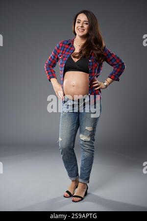 Donna incinta sicura in jeans casual e camicia aperta a quadri, sorridente e in posa con la mano sull'anca, isolata su sfondo grigio Foto Stock