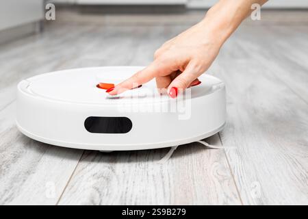 una donna preme il pulsante di accensione di un aspirapolvere robot. la casalinga accende l'aspirapolvere. Avvio dell'aspirapolvere robot. Foto Stock