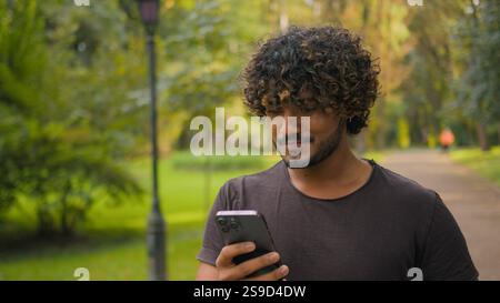 Arabo musulmano indiano ragazzo che cammina con smartphone nel parco etnico uomo assetato bere acqua da bottiglia sport mattina corsa all'aperto felice sorriso Foto Stock