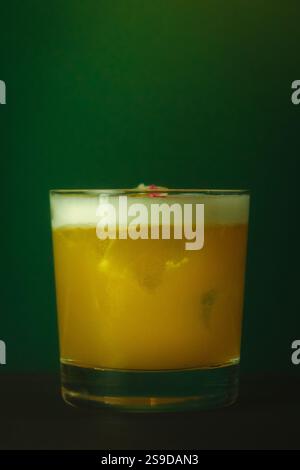 Cocktail giallo vibrante con schiuma in vetro Lowball rinfrescante bevanda su un lussureggiante sfondo verde Foto Stock
