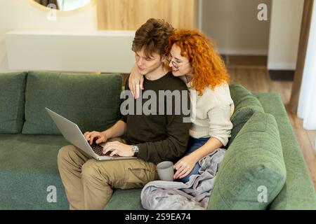 In un salotto elegante, una coppia condivide un momento caldo mentre si siedono su un divano verde e lavorano su un notebook. La luce del sole li bagna, perfetto per un rel Foto Stock
