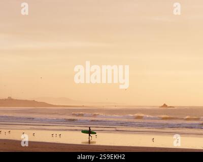 Il surfista esce dal mare con la sua tavola da surf mentre le onde si infrangono con una piccola formazione rocciosa alle spalle nella città di Essaouira, in Marocco. 25 gennaio 2025 Foto Stock