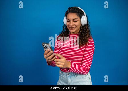 Giovane donna felice che indossa le cuffie e utilizza il telefono cellulare su sfondo blu Foto Stock