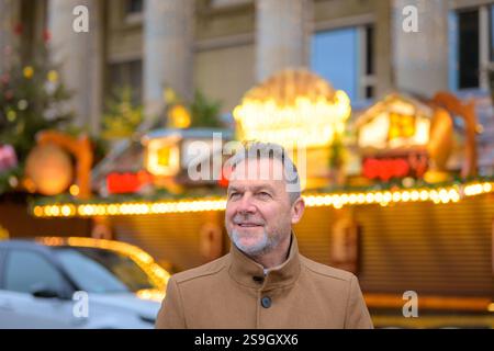 Un uomo sorridente con un cappotto beige guarda con attenzione verso l'alto, davanti a un mercatino di Natale illuminato Foto Stock