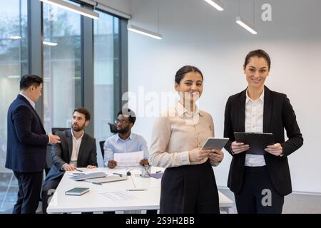 Ritratto di un gruppo di persone, due donne d'affari di successo che sorridono e guardano la telecamera, dipendenti donne all'interno dell'ufficio sul posto di lavoro in piedi in una sala riunioni. Foto Stock