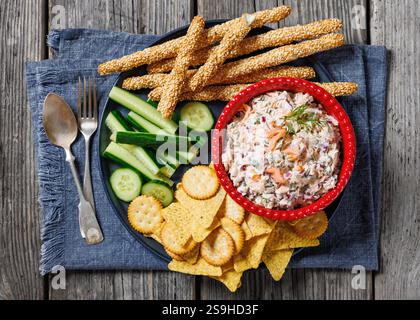 salsa di salmone con cappero, cipolla rossa, aneto fresco e panna acida nel piatto rosso con cracker, tortilla chips, cetriolo e grissini su legno rustico Foto Stock