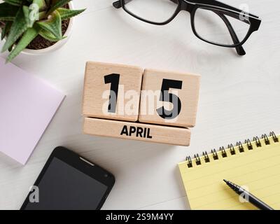 Scrivania che mostra il 15 aprile su calendario perpetuo in legno, accanto a blocco note, penna, occhiali, smartphone e impianto, ideale per promemoria fiscali Foto Stock