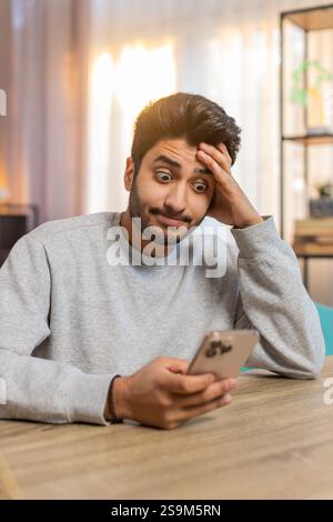 Giovane indiano seduto sul divano a tavola, scrivendo attivamente sullo smartphone, appoggiando la testa a mano nel soggiorno di casa. L'ispanico sembra concentrato, profondamente impegnato in conversazione o attività. Foto Stock