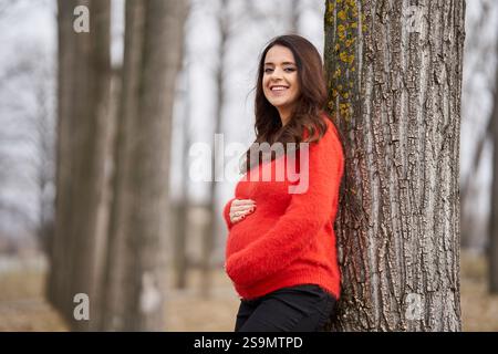 Una donna incinta con un maglione rosso e un cappello nero appoggiato contro un albero in un parco, colpita con una messa a fuoco selettiva Foto Stock