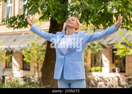 Donna d'affari matura in tuta formale che meditava a occhi chiusi in piedi sulla strada della città. Una donna di mezza età tranquilla con le braccia aperte riposa e sente la tranquillità. La donna si rilassa per alleviare lo stress. Foto Stock