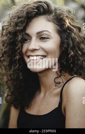 Una donna sorridente con capelli ricci gode il momento circondato dalla natura Foto Stock