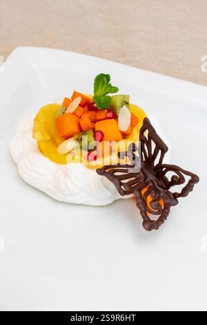 Un delizioso dessert con un mix di frutta fresca tra cui mango, kiwi e papaya su una base cremosa. Una farfalla decorativa al cioccolato e una foglia di menta Foto Stock