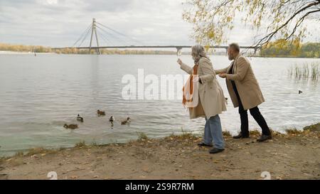 Coppia senior uomo vicino al lago che lancia il pane che dà da mangiare alle anatre insieme famiglia di mezza età passeggiata romantica nel parco autunnale all'aperto nonno in pensione Foto Stock