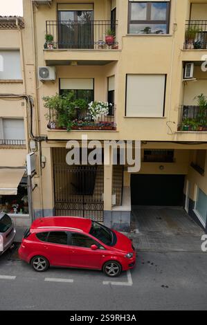 Una strada cittadina presenta una piccola facciata residenziale con balconi e un'auto rossa parcheggiata sotto. Le piante decorano i balconi, creando un Foto Stock