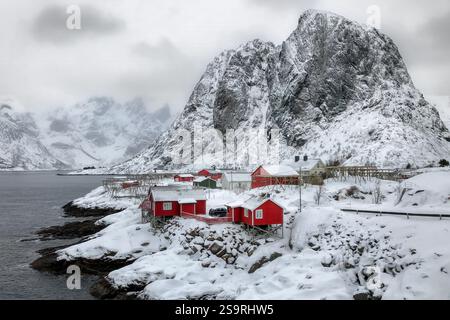 Cabine rosse ad Harmony, Norvegia, sedetevi accoccolati lungo una costa innevata sotto torreggianti e aspre montagne. I cieli ricoperti suggeriscono a basse temperature, con un Foto Stock