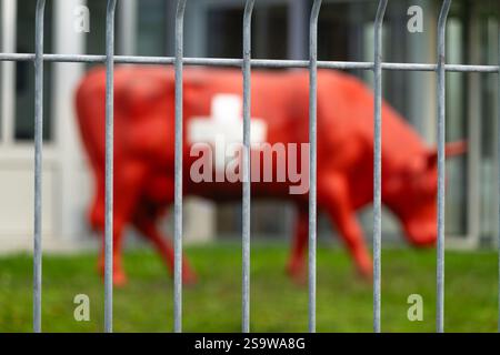 Una scultura di mucca rossa sfocata nei colori della bandiera svizzera è visibile attraverso una recinzione metallica. La scultura presenta un simbolo a croce bianca. Lo sfondo suggerisce Foto Stock