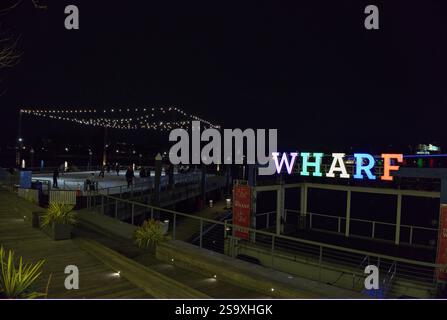 Foto notturna del cartello al neon del Wharf con pista di pattinaggio sullo sfondo. Il quartiere offre eventi musicali, ristoranti, negozi, ecc. Foto Stock