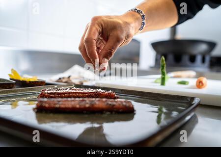 Primo piano di uno chef che condita carne gourmet su un vassoio, con verdure come asparagi e carote sullo sfondo. Foto Stock
