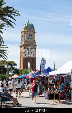 Le persone passeranno davanti alle bancarelle del mercato domenicale accanto alla Torre dell'orologio Carlo Catani Memorial su un'assolata St Kilda Esplanade, Melbourne Foto Stock