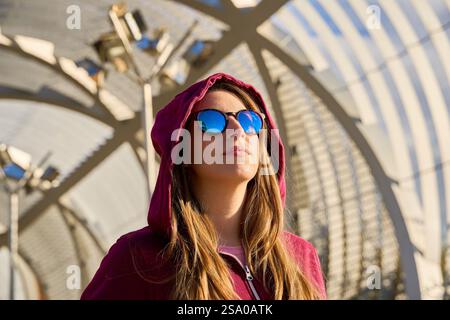 Ritratto di una giovane donna alla moda che indossa una felpa con cappuccio e occhiali da sole a specchio in un ambiente urbano moderno Foto Stock