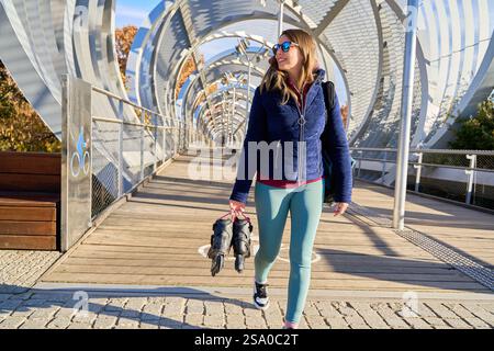 Donna sportiva che porta pattini a rotelle mentre cammina lungo un moderno ponte cittadino, goditi la giornata di sole e abbraccia uno stile di vita attivo Foto Stock