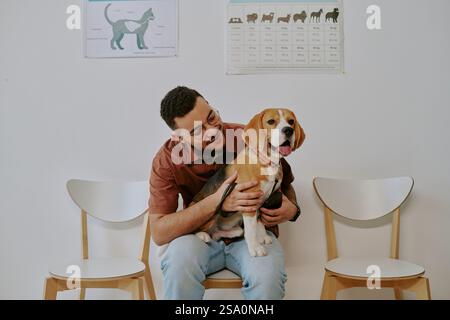 Giovane sorridente mentre tiene un beagle in grembo, seduto su una sedia di fronte al muro bianco con poster educativi appesi dietro di loro Foto Stock