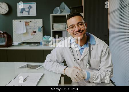Medico sorridente seduto alla scrivania con attrezzature mediche e documenti intorno, mostrando un comportamento amichevole e professionale in un ambiente ben organizzato di ufficio medico Foto Stock
