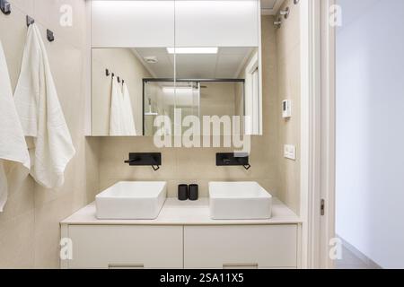 Un bagno contemporaneo con doppio lavandino, eleganti rubinetti neri, un grande specchio, piastrelle neutre e morbidi asciugamani bianchi. Foto Stock