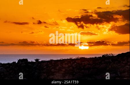 Uno splendido tramonto sull'oceano, con vivaci sfumature arancioni e gialle che riempiono il cielo. Le nuvole sagomate aggiungono profondità alla scena, mentre l'orizzonte si ripete Foto Stock