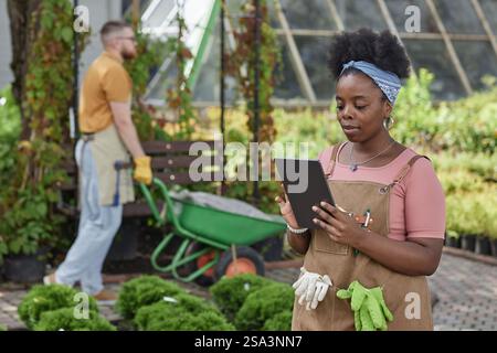 Colpo medio di calma afroamericana orticulturalista femminile in grembiule utilizzando computer tablet, ispezionando arbusti e fiori per garantire piante sane e prive di malattie nel centro giardino Foto Stock