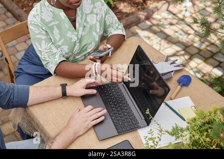 Vista dall'alto dell'irriconoscibile architetto paesaggista afroamericano che punta sullo schermo di un laptop discutendo la progettazione dello spazio pubblico con un collega, mentre l'uomo tiene le dita sul touchpad al tavolo all'aperto Foto Stock