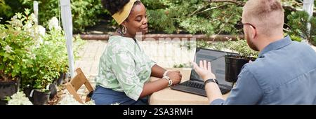 Immagine di intestazione del sito web di una donna sorridente paesaggista di etnia nera seduta a tavola con un collega maschio che discute del progetto del giardino, scegliendo arbusti e alberi nel vivaio vegetale Foto Stock