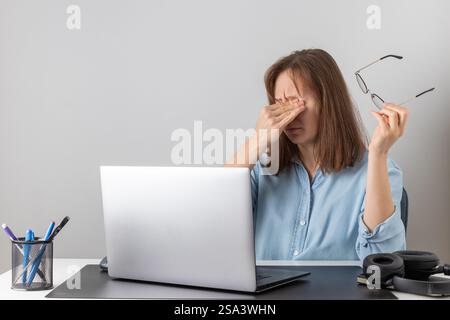 Una freelance stressata a una scrivania, che si sfrega gli occhi mentre lavora su un notebook. Indossa gli occhiali, sembra esausta e lotta con gli strai degli occhi Foto Stock