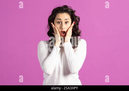 Concetto di stile di vita. Bella donna bruna con espressione facciale eccitata che tiene le mani sulle guance del viso e guarda la fotocamera a bocca aperta. Pur Foto Stock