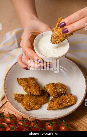 Una donna che immerge un pepite dorato croccante in una cremosa salsa bianca. Un piatto con più pepite e pomodori ciliegini freschi su un asse di legno. Toni caldi AN Foto Stock