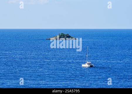 Uno yacht catamarano naviga verso una piccola isola astratta solitaria disabitata nel mare blu Foto Stock